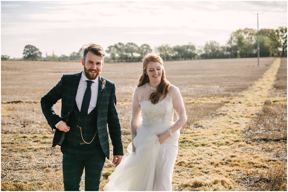 Couple walking in a cornfield at Curradine Barns; Groom with beard and tartan suit