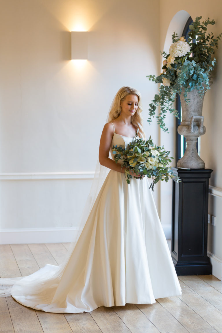 Bride wearing classic A line wedding dress, holding bouquet of white flowers with green foliage, in front of windows in the orangery at Alrewas Hayes