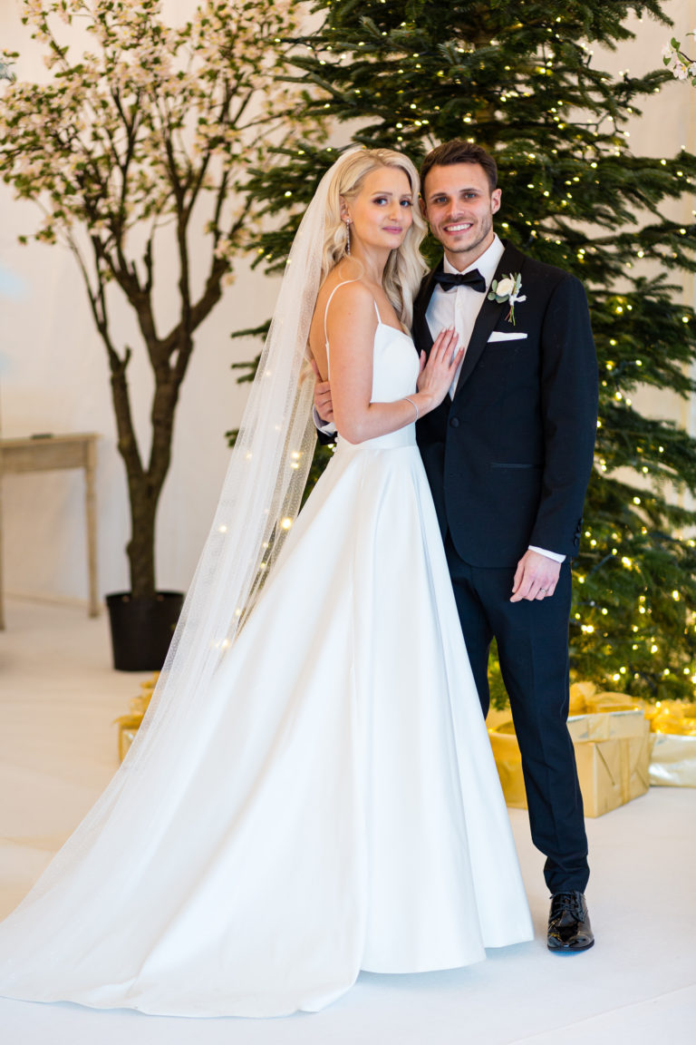 Bride and Groom smiling in front of a large Christmas tree inside the marquee at their winter wedding at Alrewas Hayes