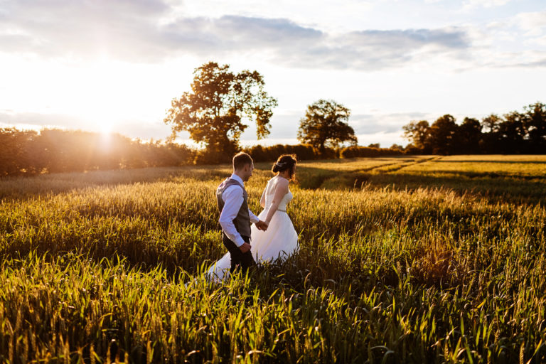 Bride holding Grooms hand walking through a cornfield at sunset at Alrewas Hayes