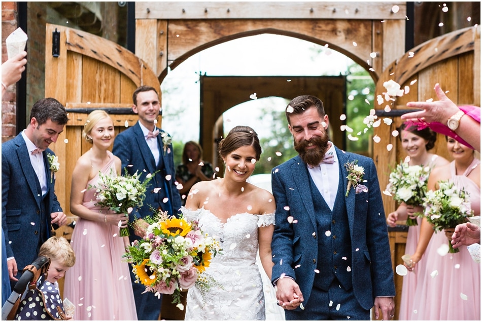 Dickie bow wearing bearded Groom with his Bride walking through confetti at Shustoke Barn