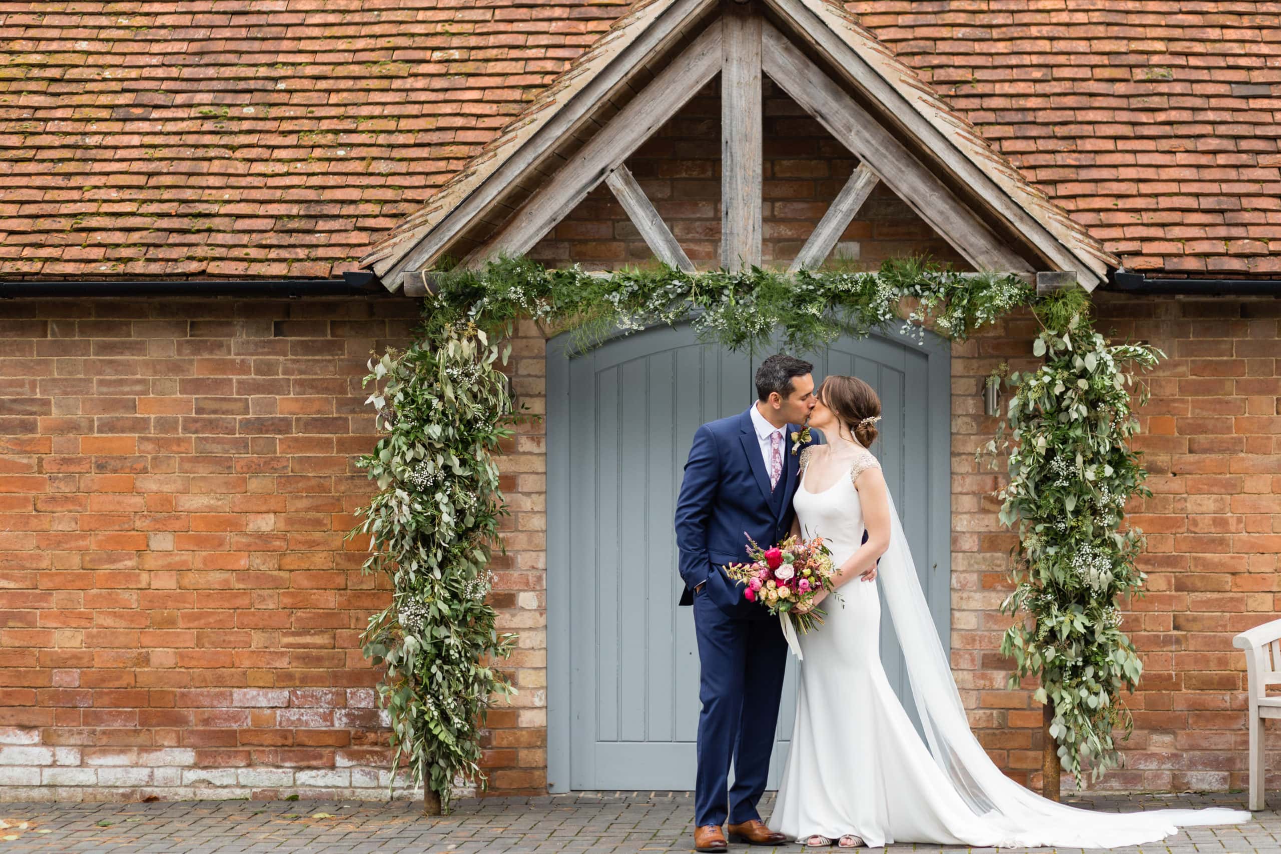 Bride and Groom kissing in under an archway of green foliage in front of grey door