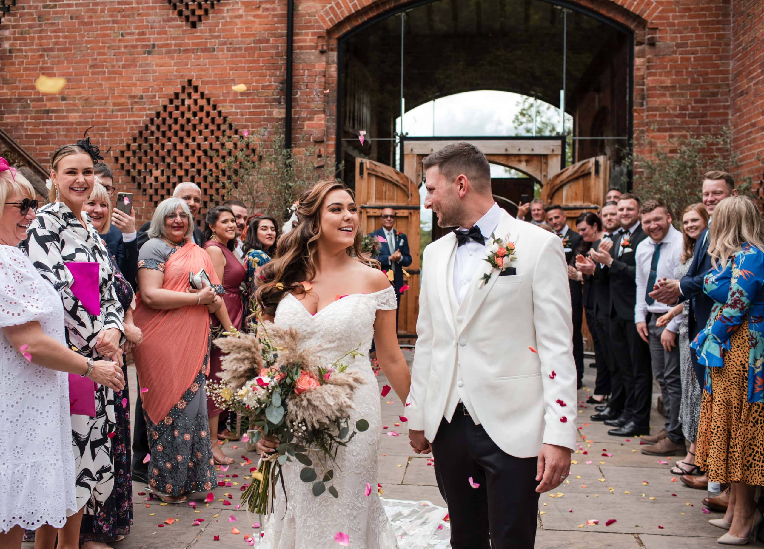 Bride holding pampas bouquet and Groom wearing white tuxedo with confetti at Shustoke Barn