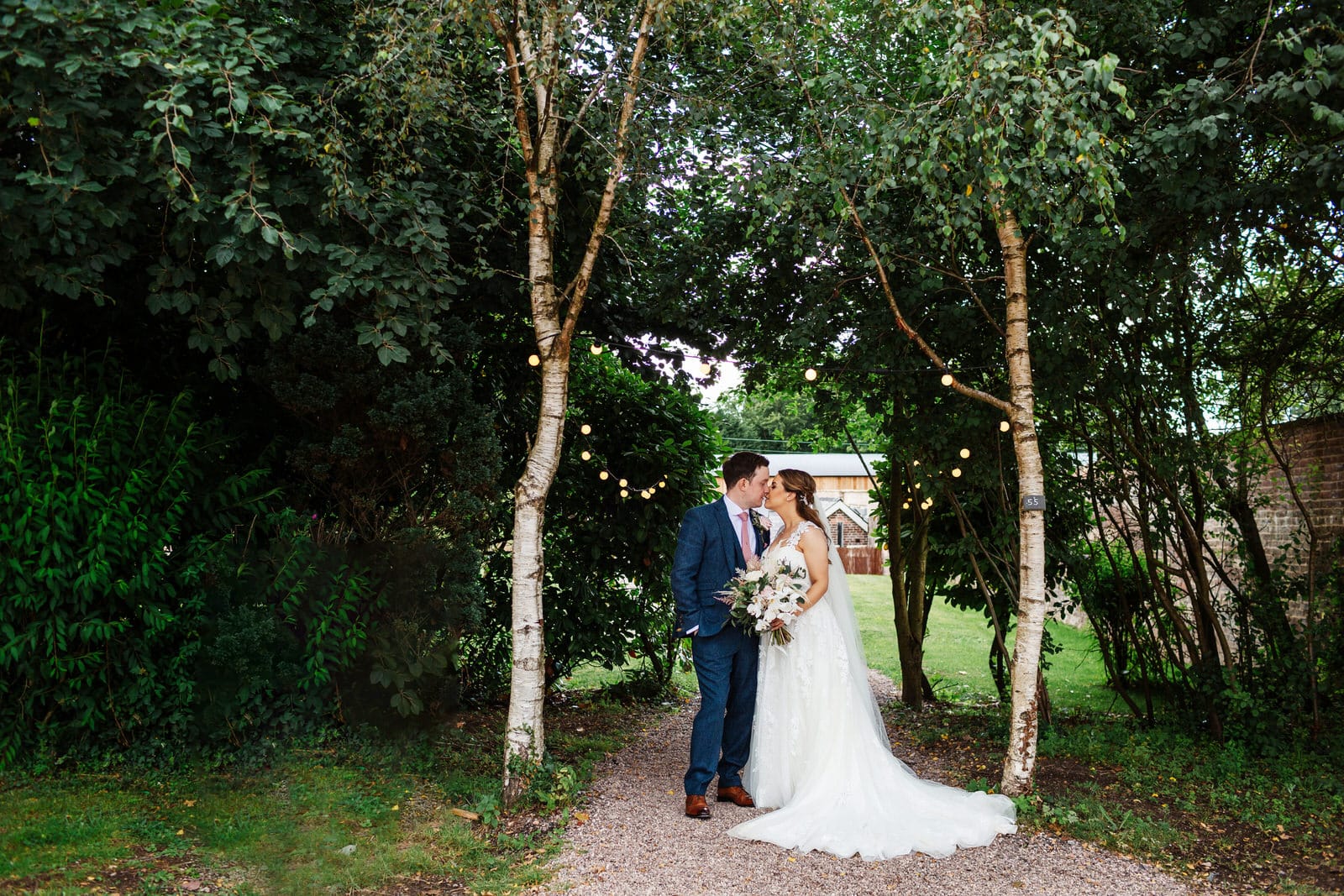 Groom wearing blue suit kisses his Bride standing in-between silver birch trees
