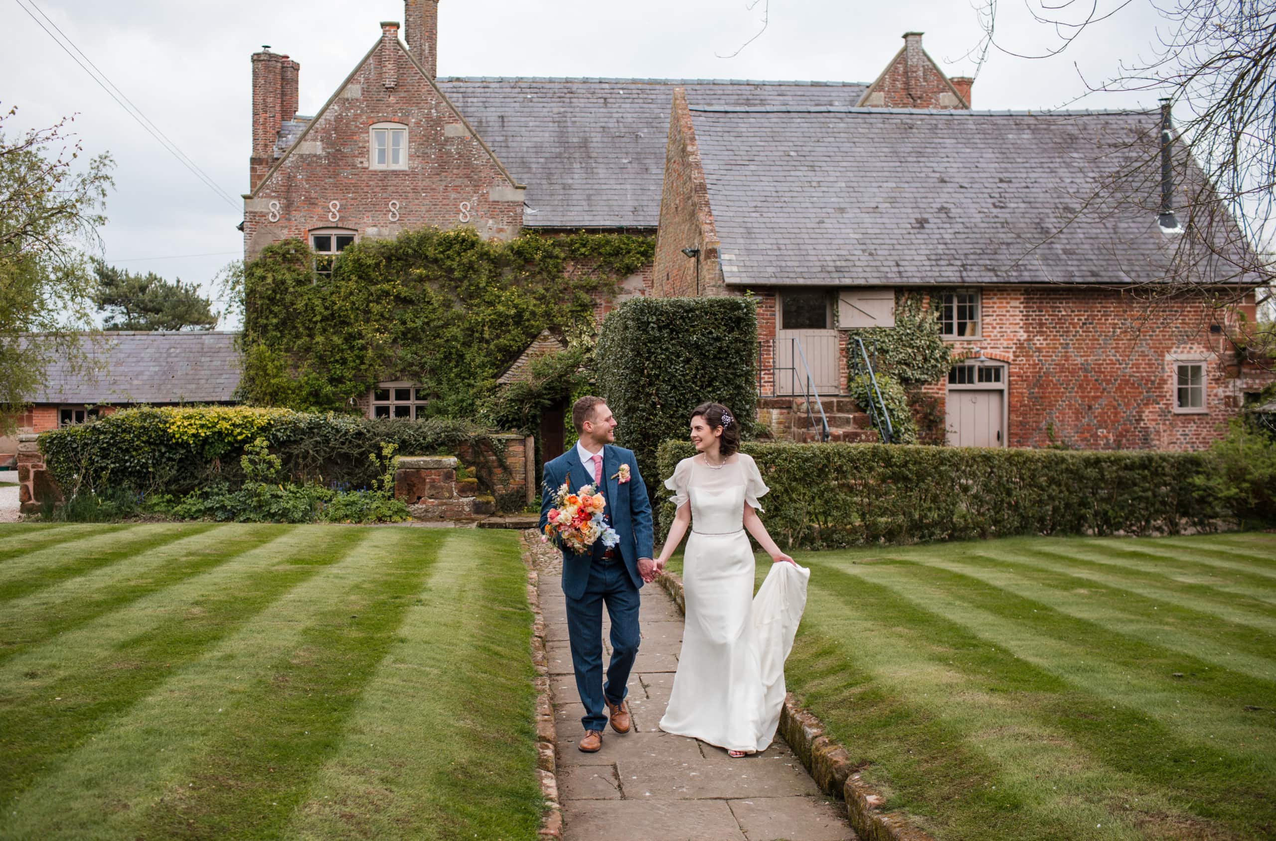 newly married couple stroll hand in hand along a path leading away from an old farmhouse