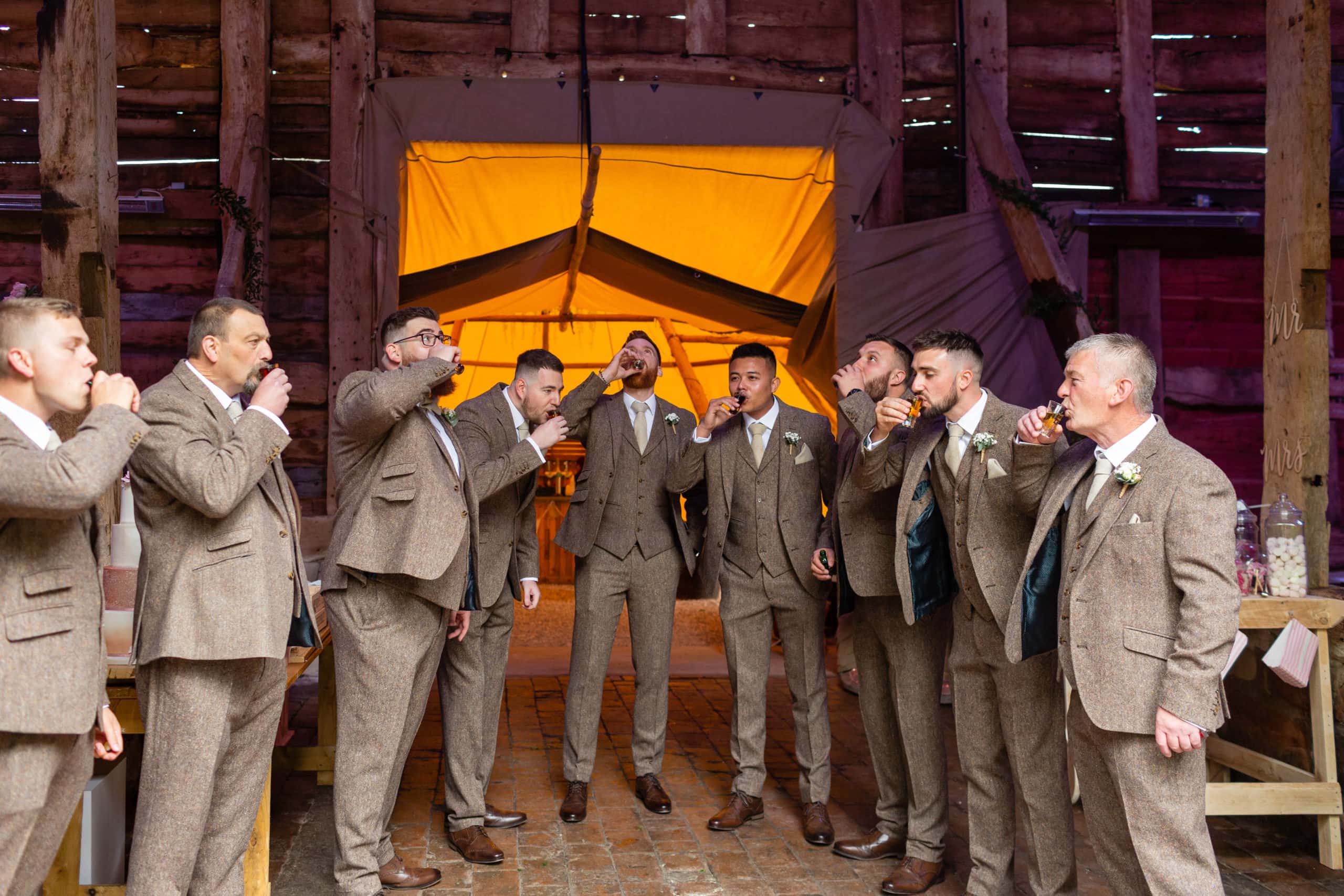 eight Groomsmen standing toghether wearing brown tweed suits drink a shot