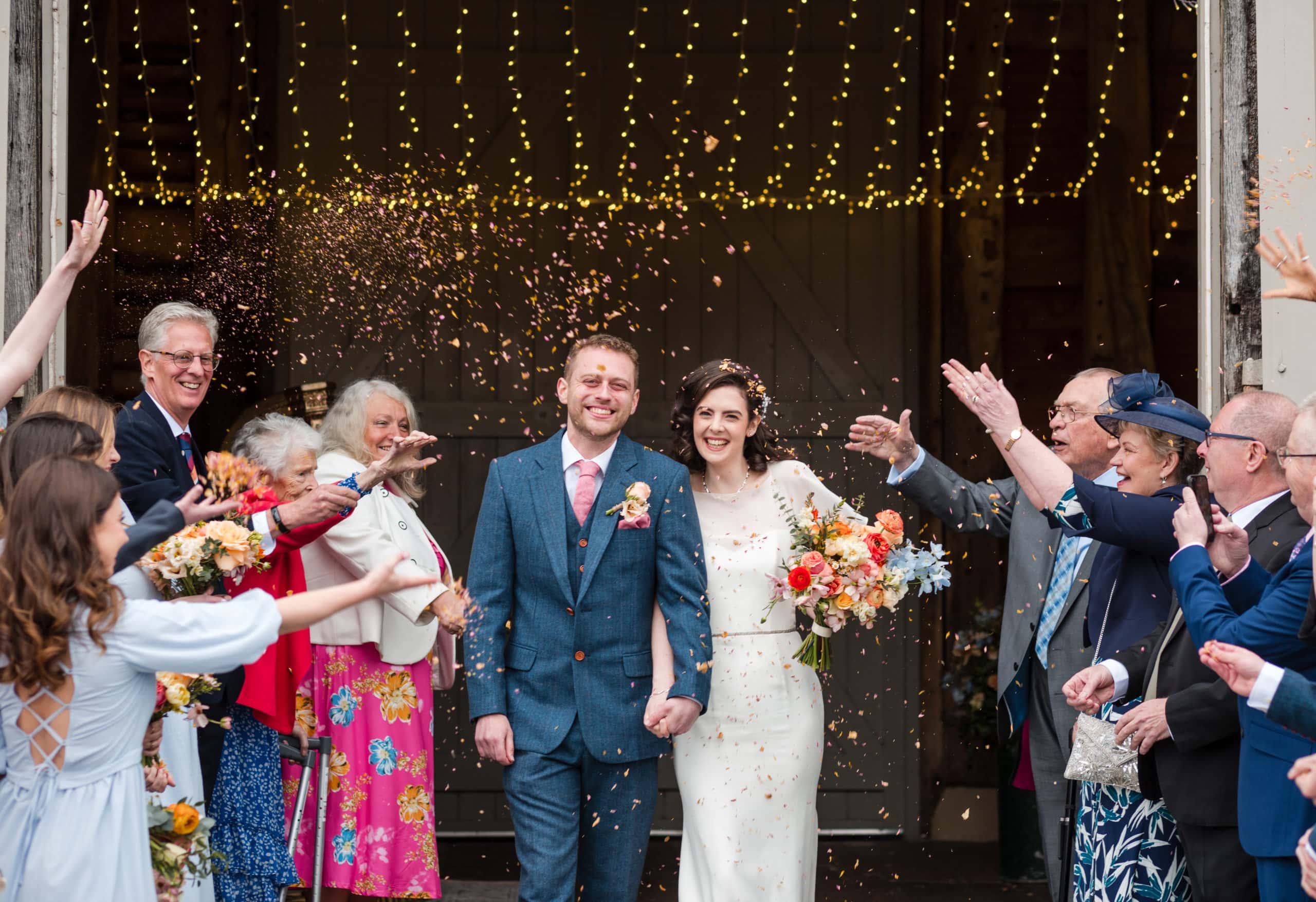 smiling Bride and Groom walk out of Pimhill Barn through confetti holding hands. Bride carries a colourful bouquet