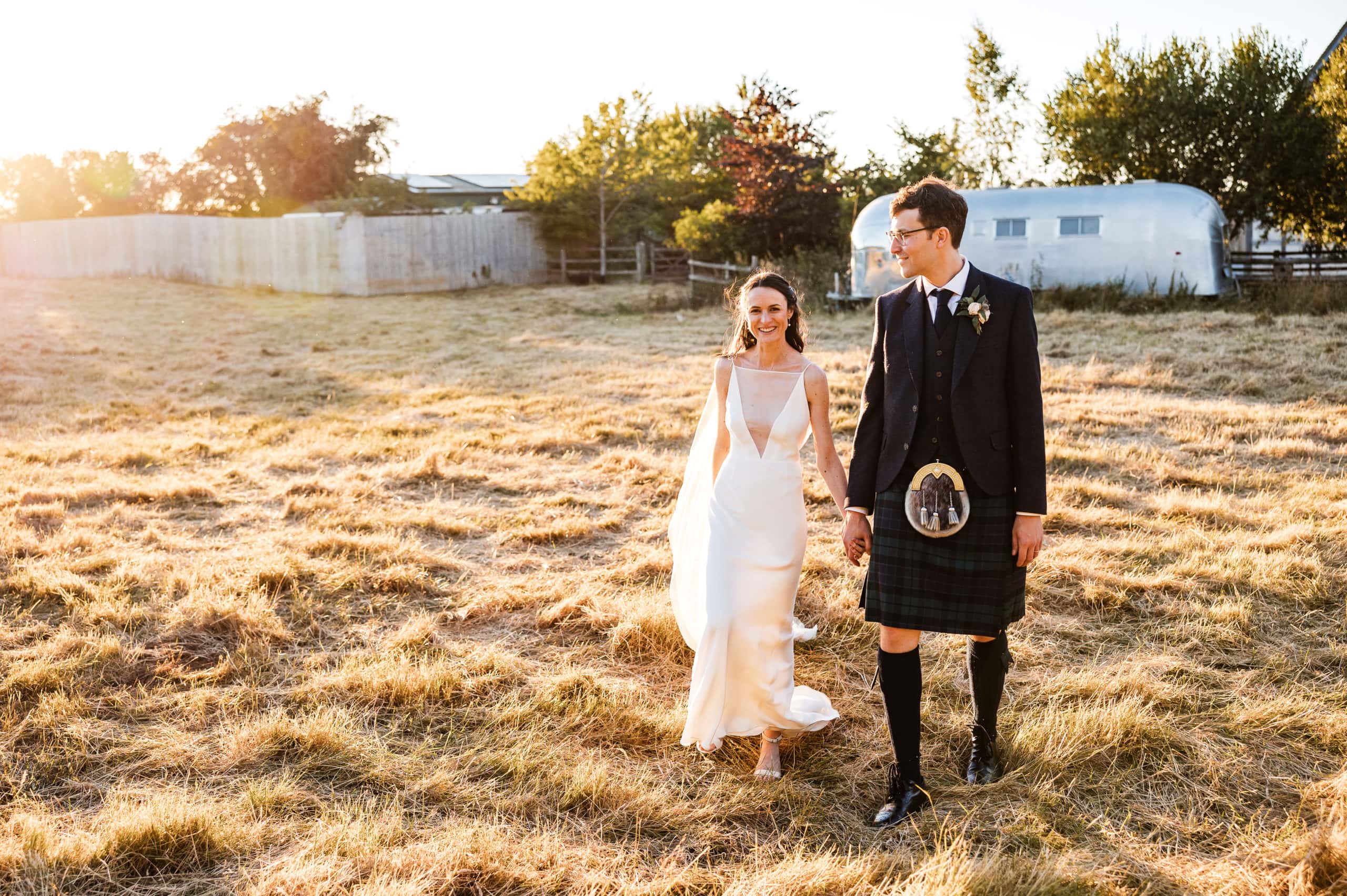 Groom wearing a kilt holding hands with bride in a hay field at sunset