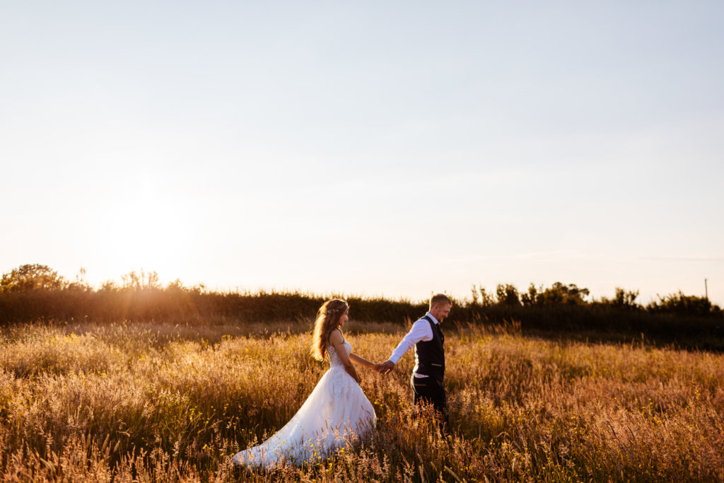 Bride and Groom walking through long grass meadow at Sunset
