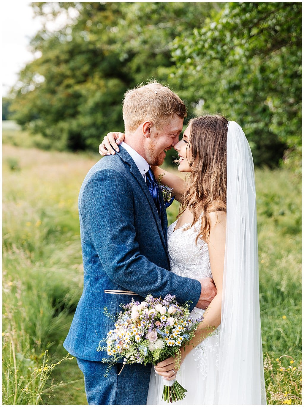 Groom wearing blue tweed suit hugs his Bride in a field in Smallwood village