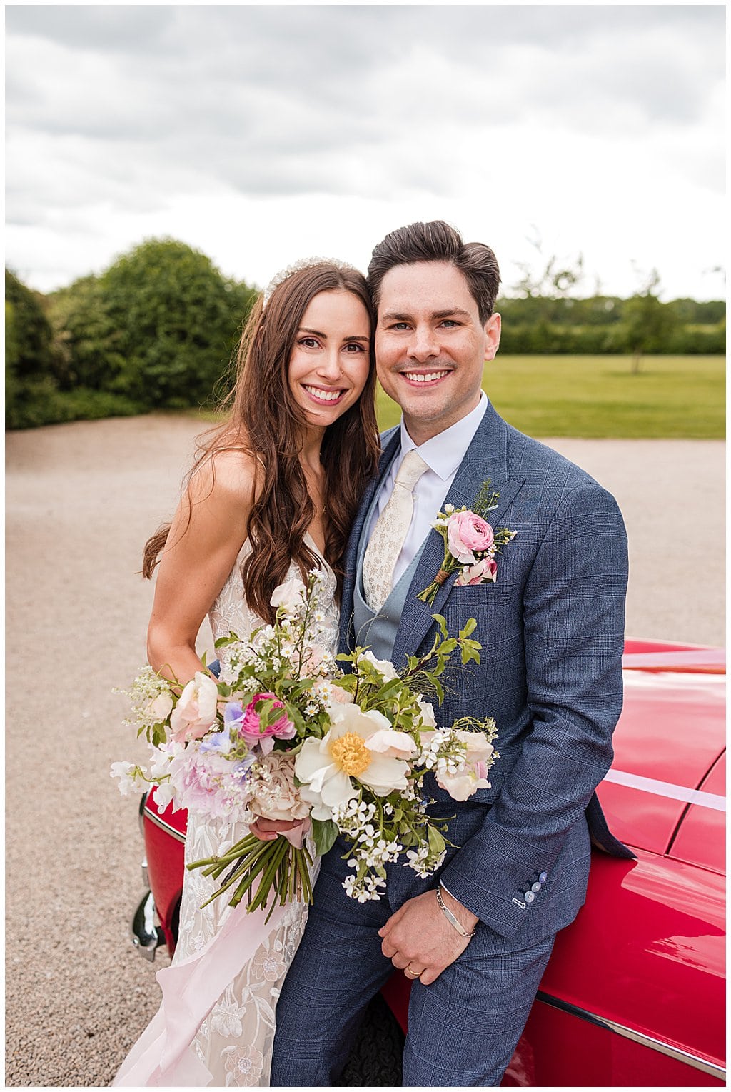 Happy portrait of a Bride and Groom sitting on a red sports car in the driveway of Alrewas Hayes. Bride wears hair loose and holds ribbon tied relaxed bouquet. Groom wears blue check three piece suit