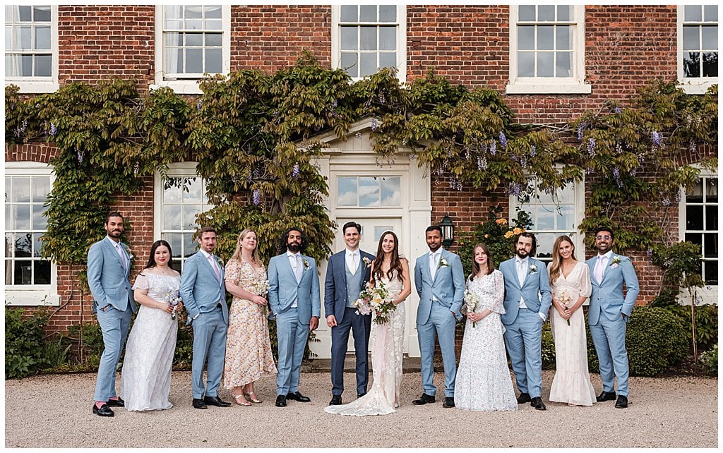 Group photo of a Bridal Party in front of a country Manor House. Groomsmen wear light blue suits and Bridesmaids mismatched floral dresses