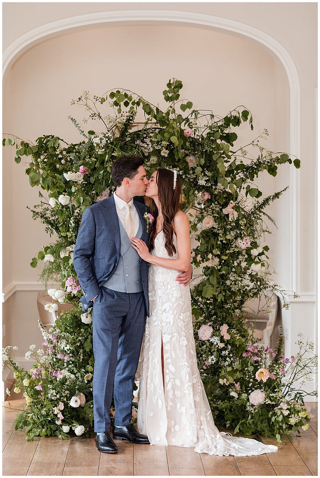 Bride wearing Hermione de Paula kissing Groom in blue three piece suit, standing in front of a floral arch backdrop in the ceremony room at Alrewas Hayes