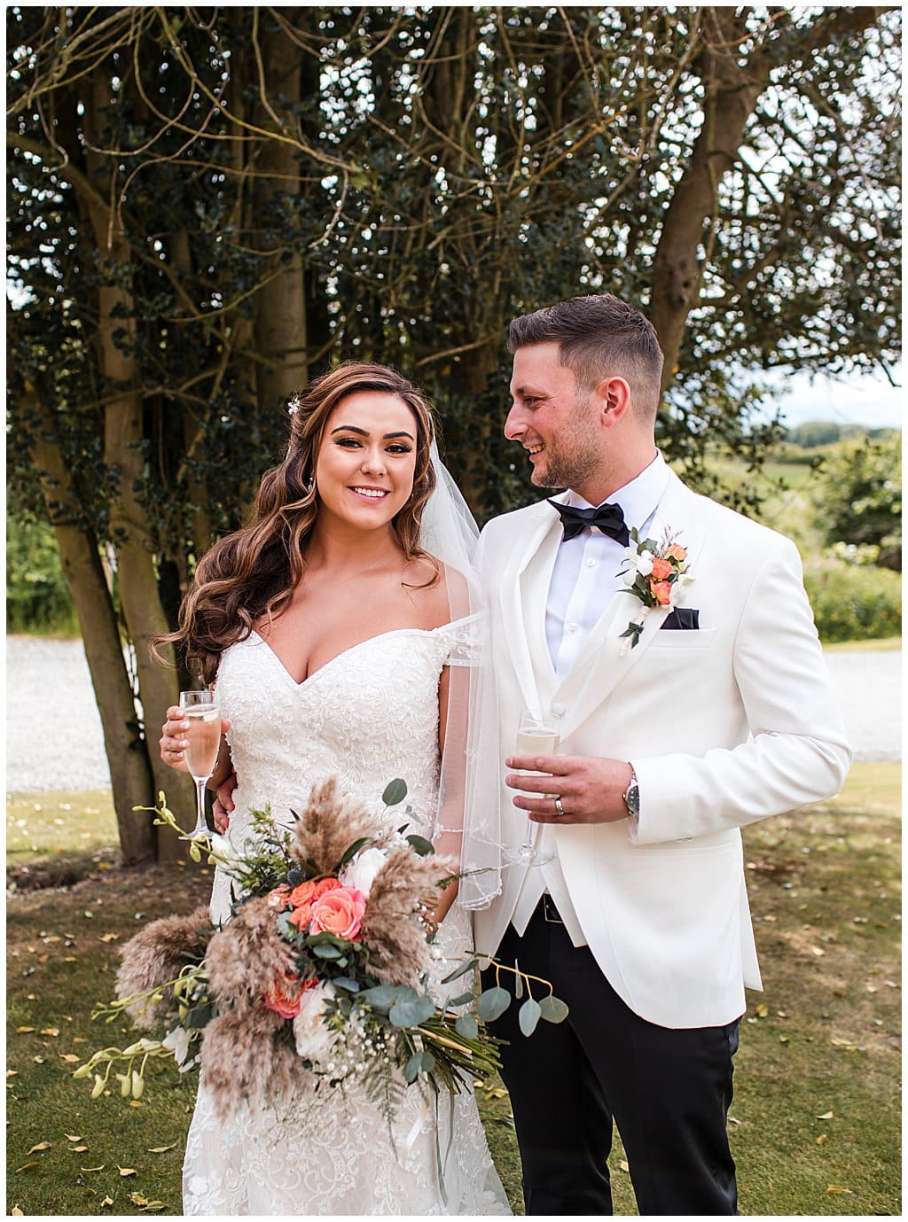 Relaxed smiley Bride and Groom holding champagne. Bride holds pampas grass bouquet. 