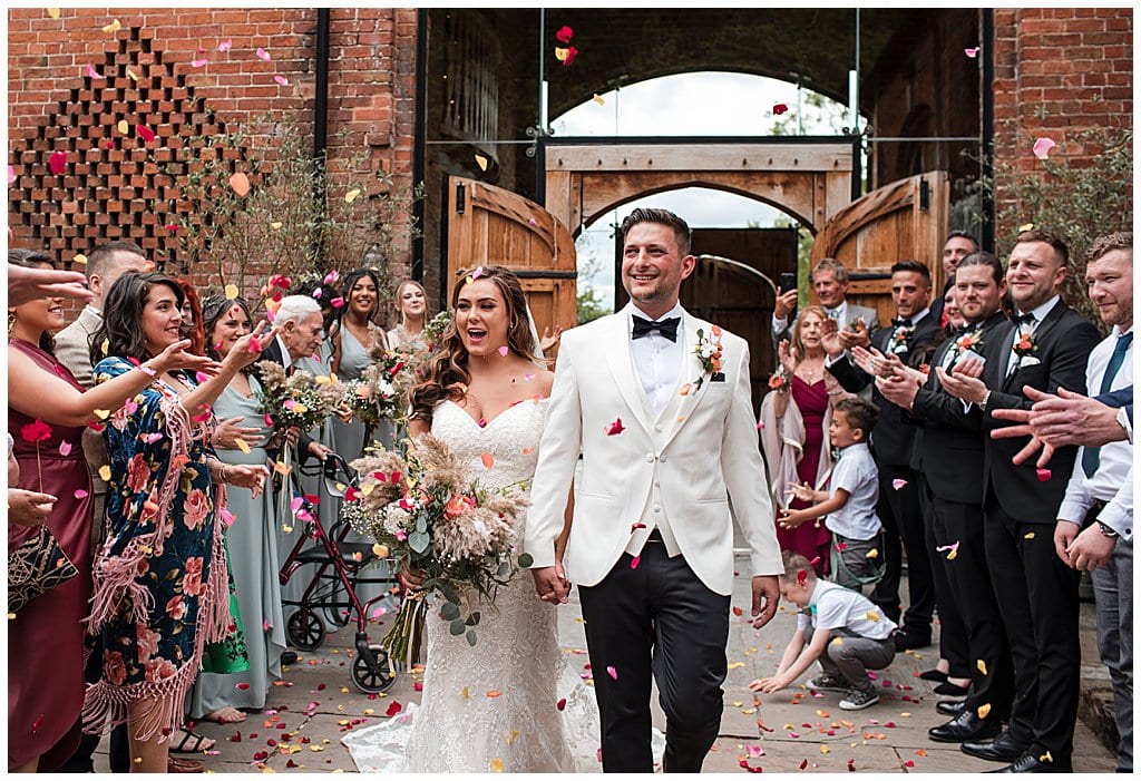 Just married couple walk out of Shustoke Barn through colourful petal confetti 