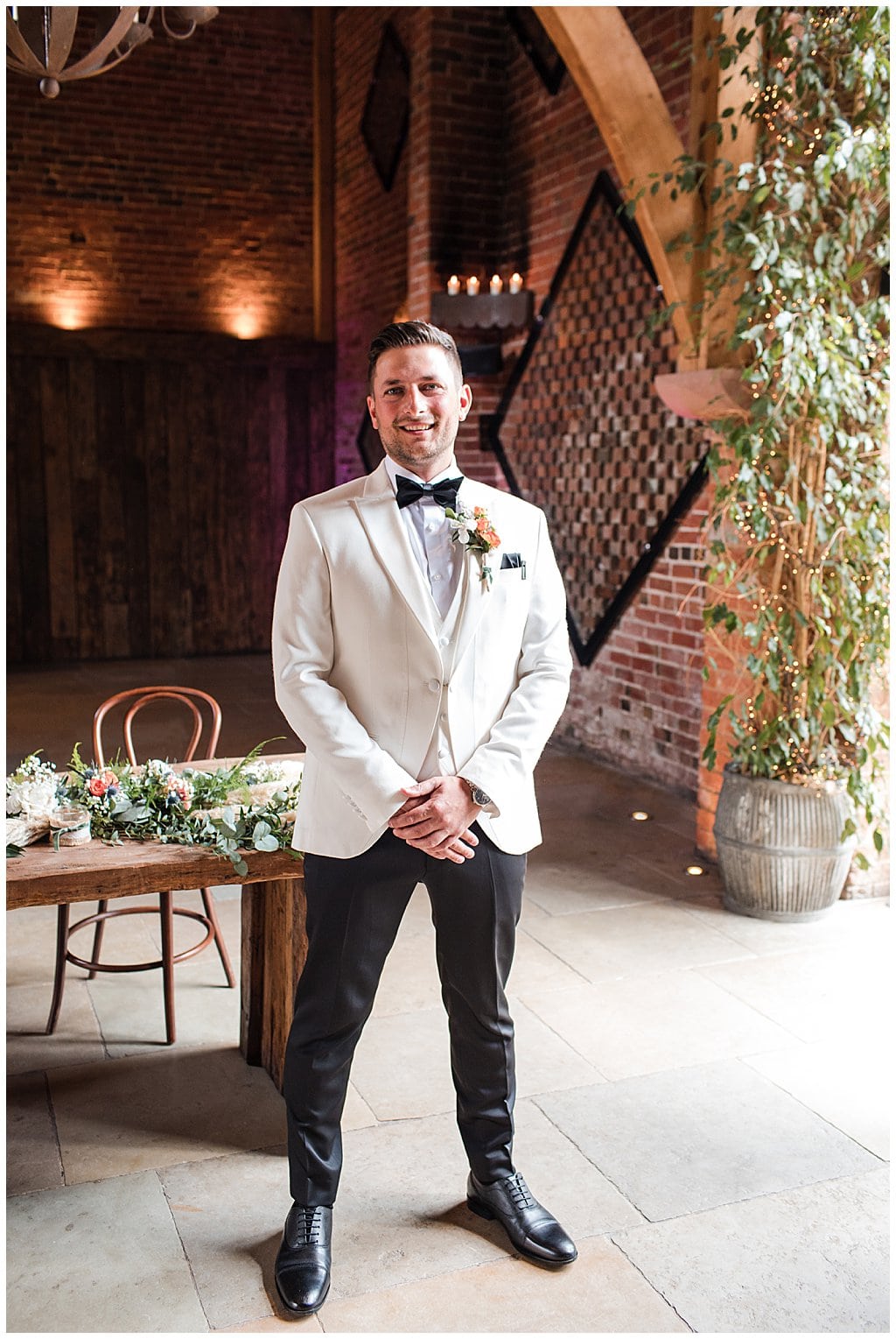 Groom wearing white tuxedo and black bow tie, stands waiting for Bride at Shustoke Barn ceremony