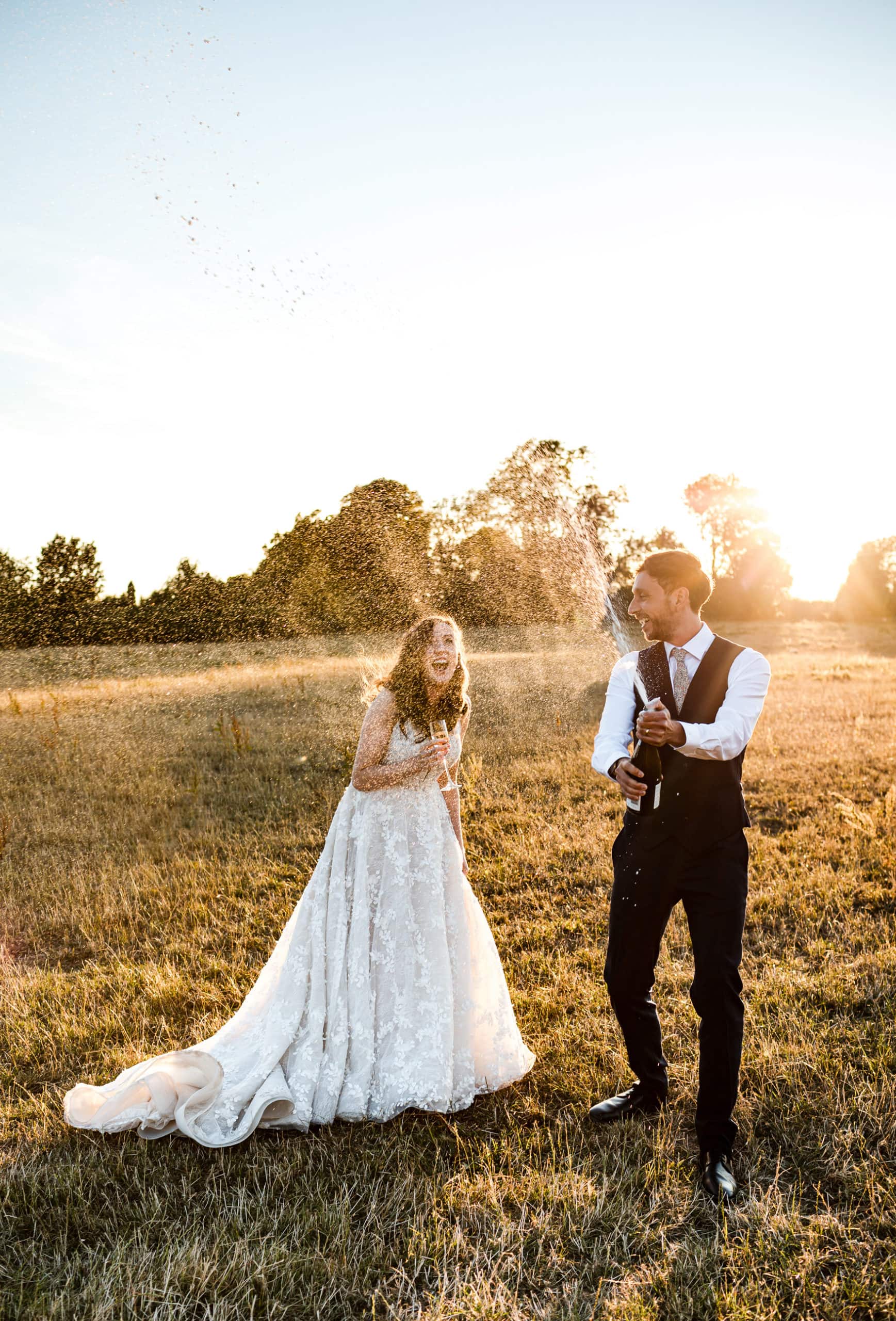 Bride and Groom spray Prosecco at sunset in fields by Curradine Barns