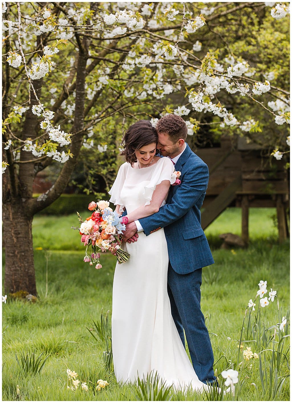 Bride and Groom cuddle in front of spring blossom trees in Pimhill Barn gardens