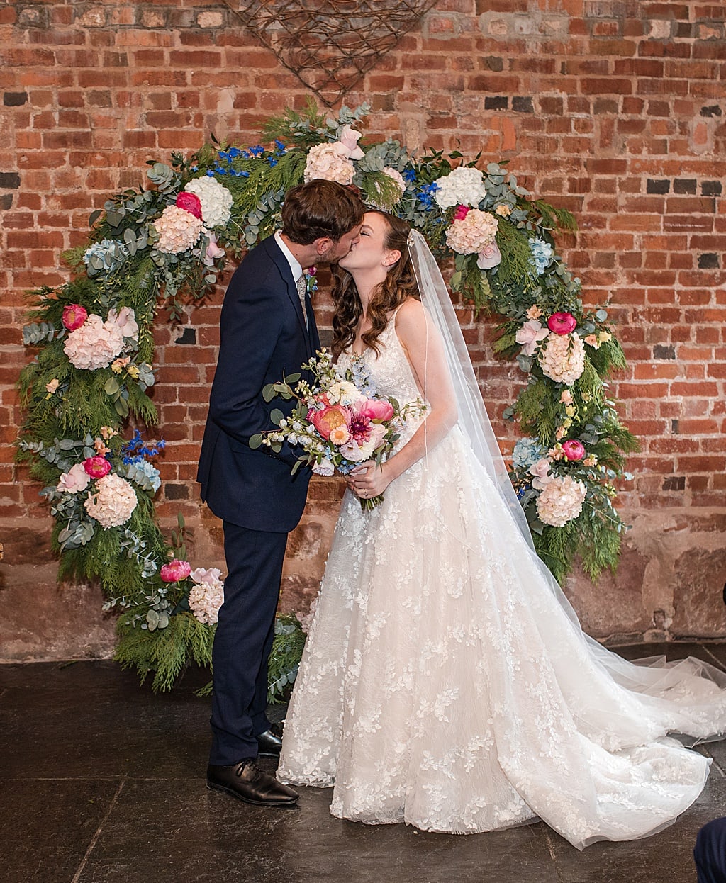 Bride and Groom share a first married kiss standing in front of a circular backdrop of flowers