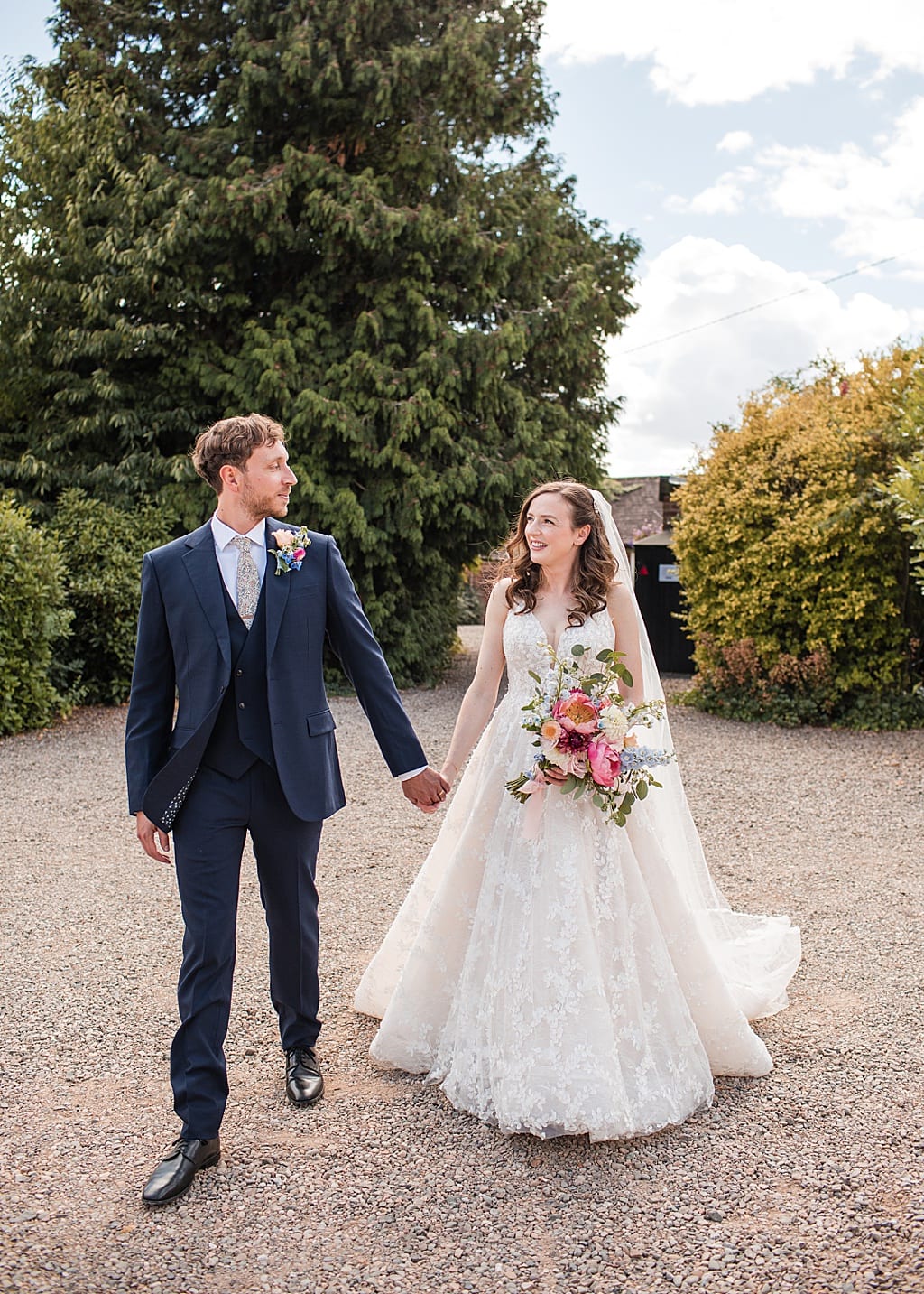 Groom wearing navy blue suit holding hands with his Bride carrying a colourful bouquet, walking together