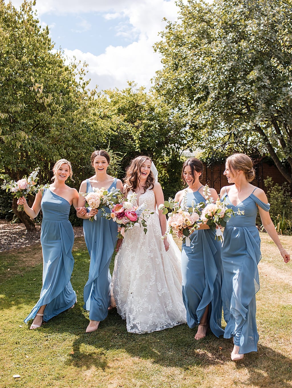 Bride and her Bridesmaids wearing blue chiffon dresses walk together laughing in the garden at Curradine Barn