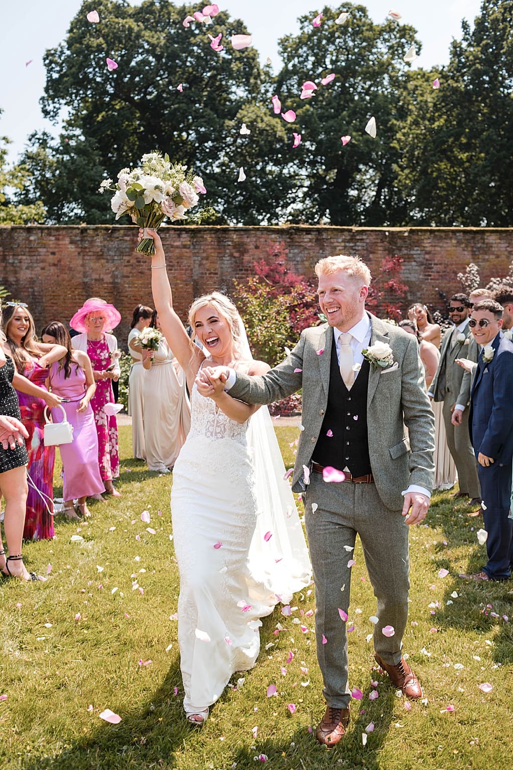 Bride holds her bouquet above her head walking through petal confetti