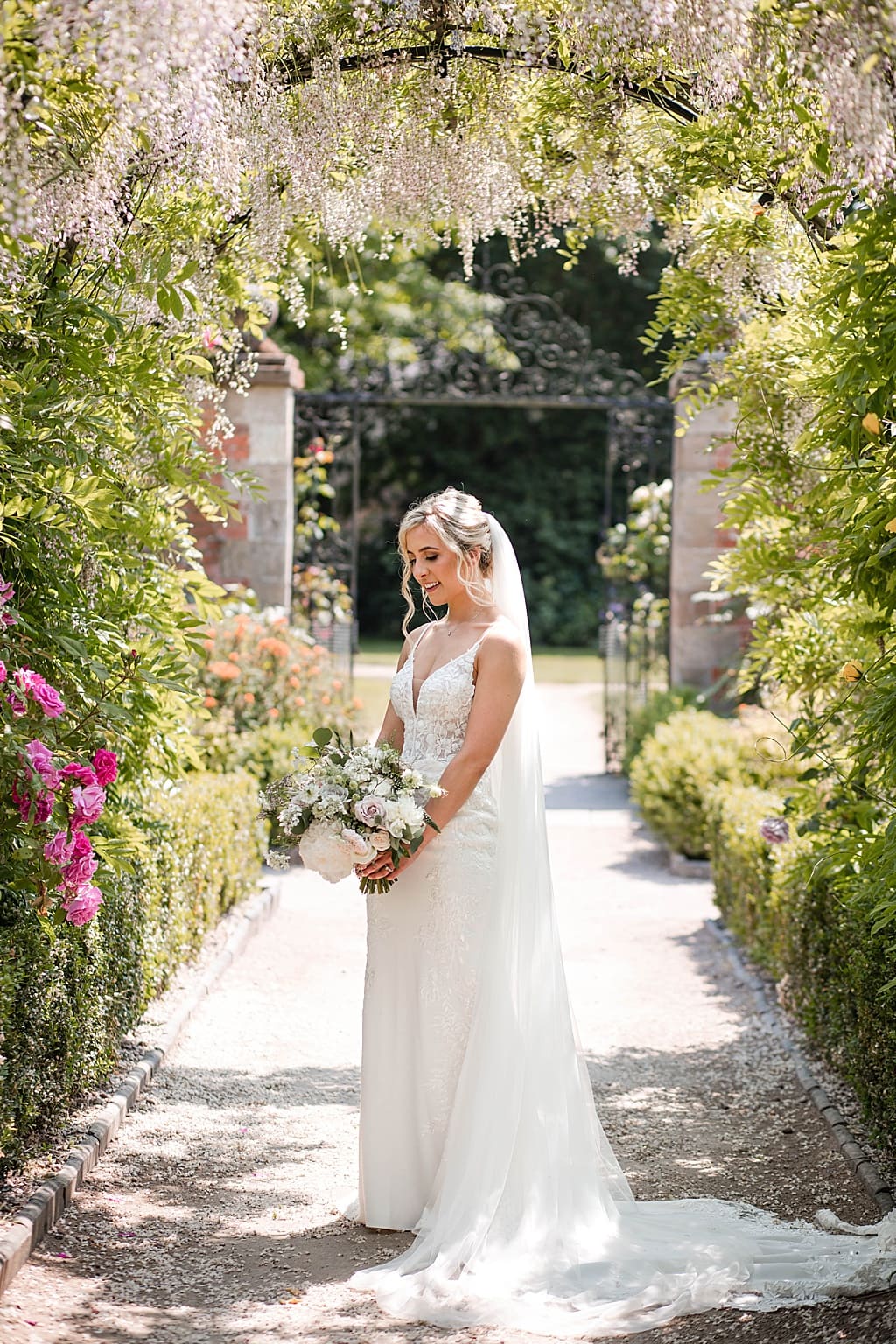 Bride stands under a wisteria arch looking at her bouquet at Thorpe Garden