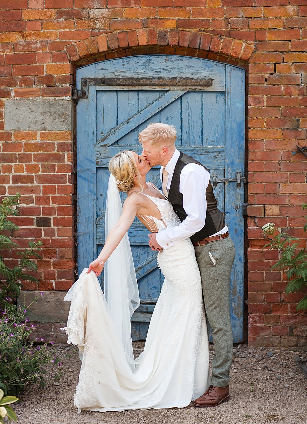 Groom wearing waistcoat and not jacket kisses his Bride in front of a blue door. Bride holds out her dress