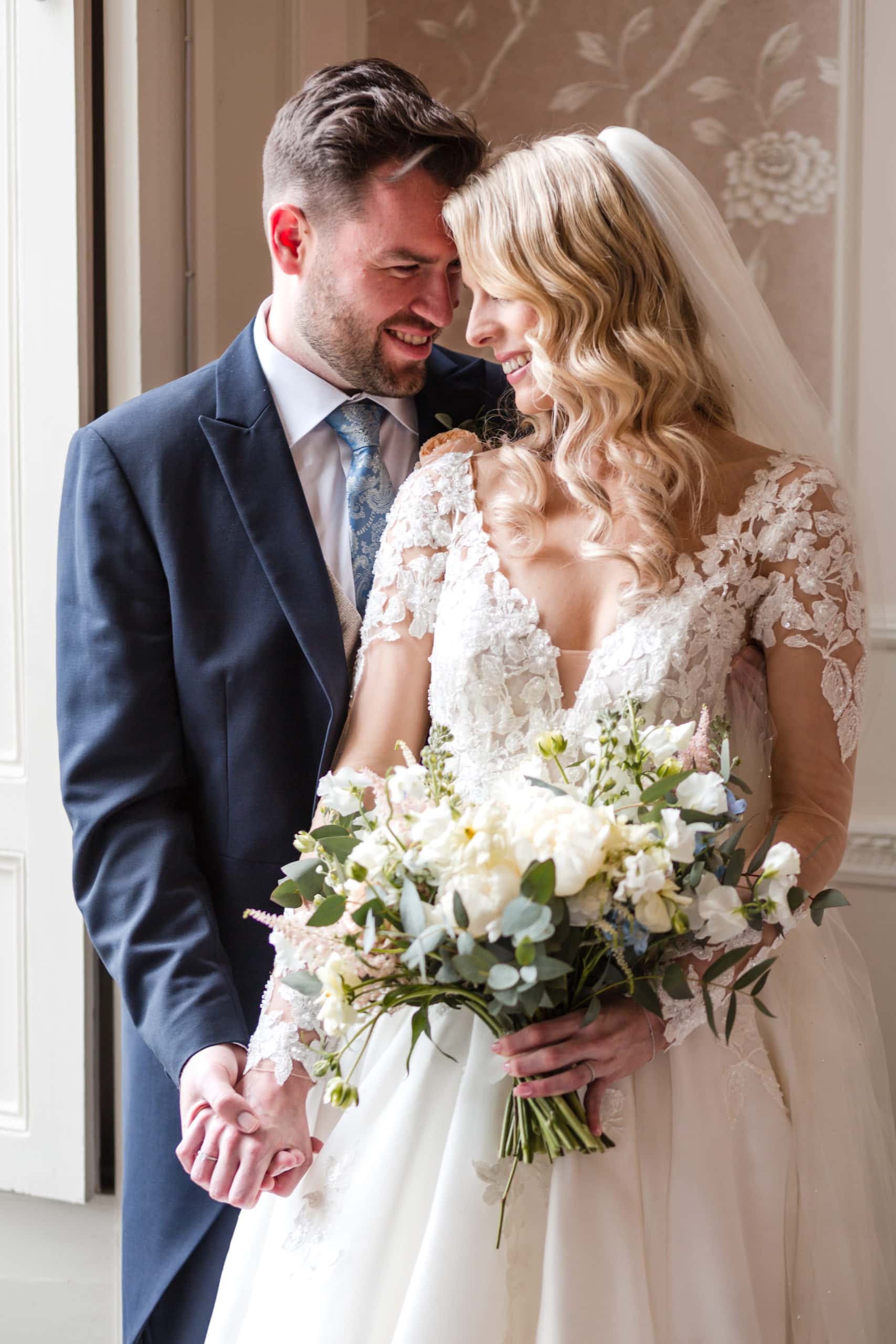 Blonde haired Bride wearing long sleeved lace dress and holding bouquet snuggles with Groom at Rowton Castle