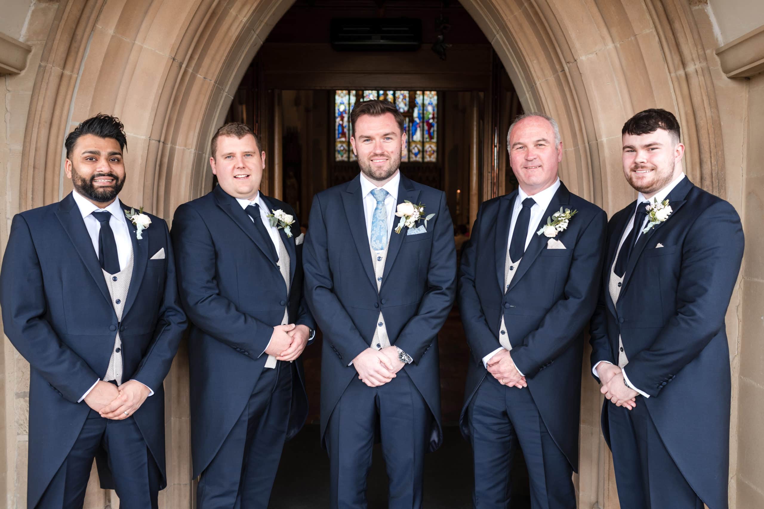 Groom and Groomsmen wearing traditional navy blue morning suits stand outside the entrance to Shrewsbury Cathedral