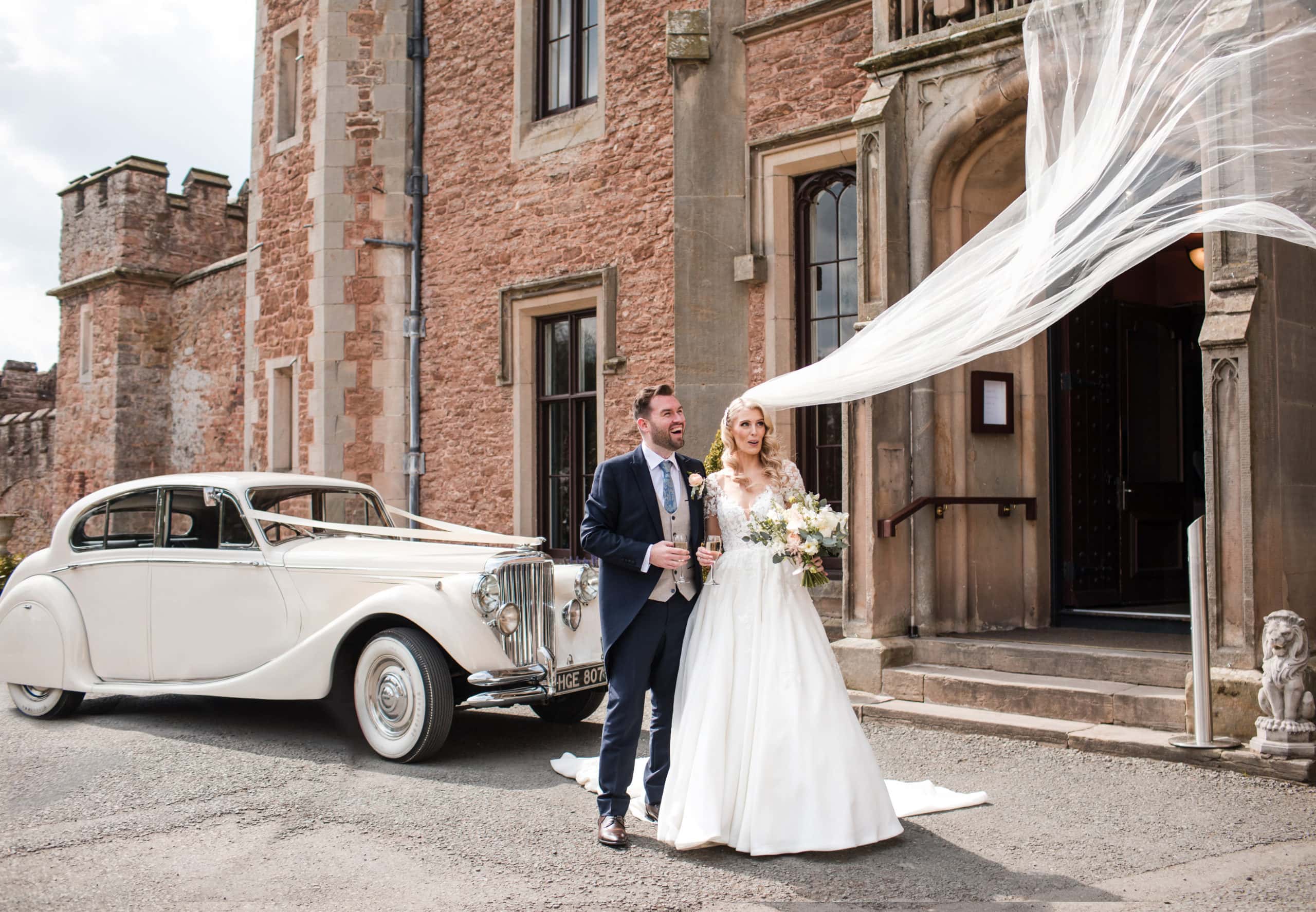 Bride and Groom stand with white vintage wedding car in front of Rowton Castle. Gust of wind blows brides veil into the air