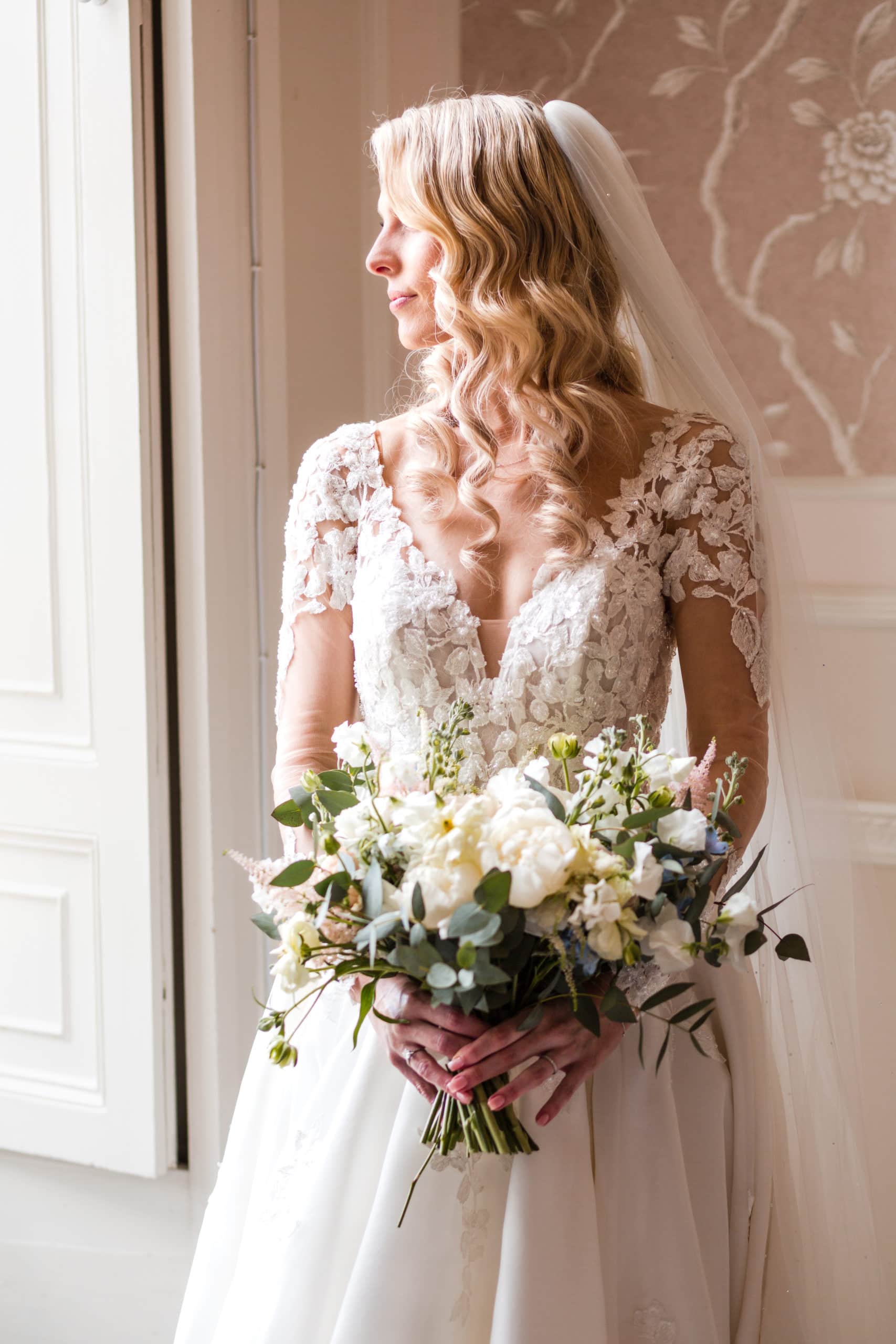 window lit portrait of a bride wearing Justin Alexander illusion sleeve wedding dress. Bride has blonde Hollywood wave hairstyle and holds white and green bouquet 