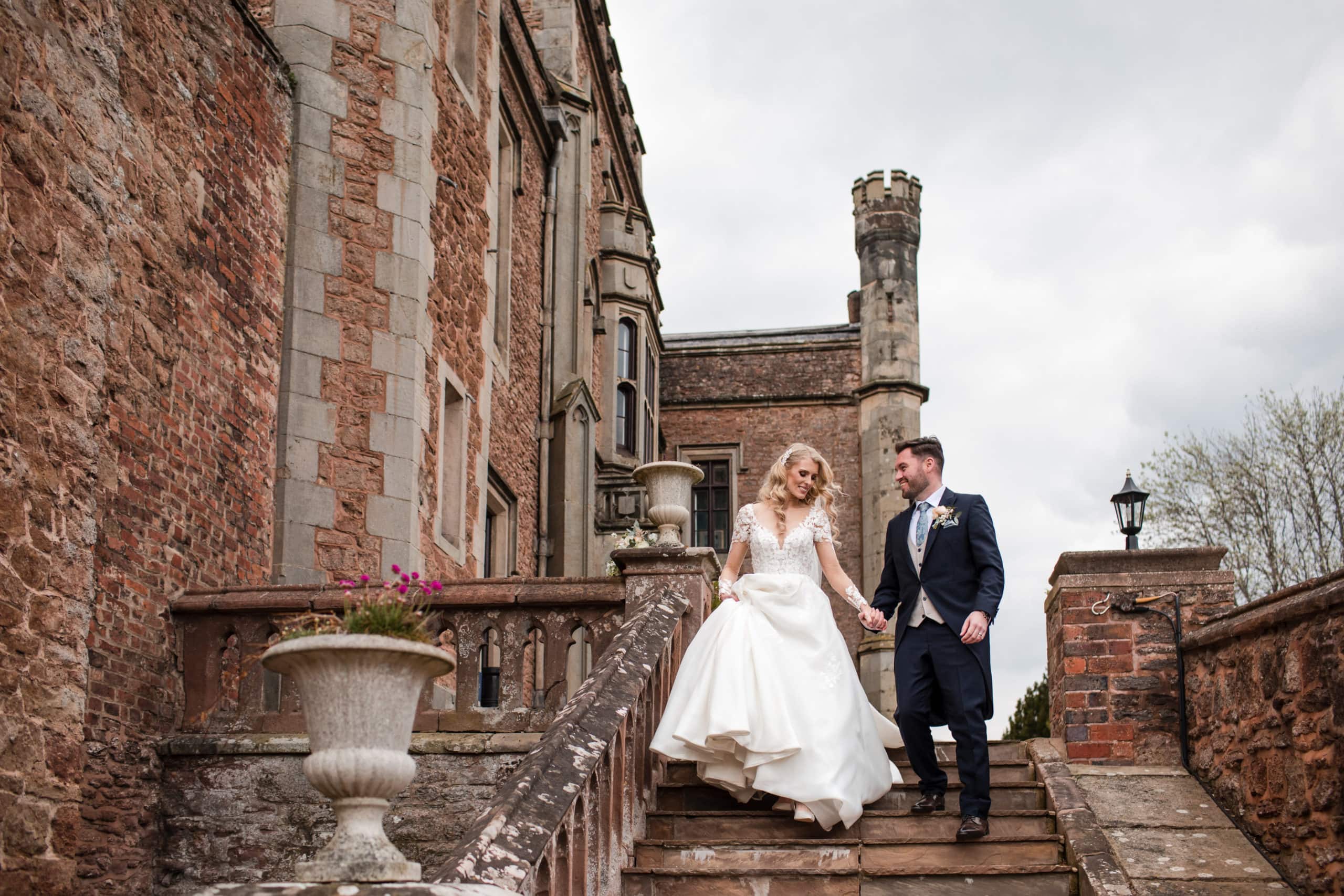 Bride holds her dress up in one hand as she walks down some steep steps at Rowton Castle, holding hand with her Groom 