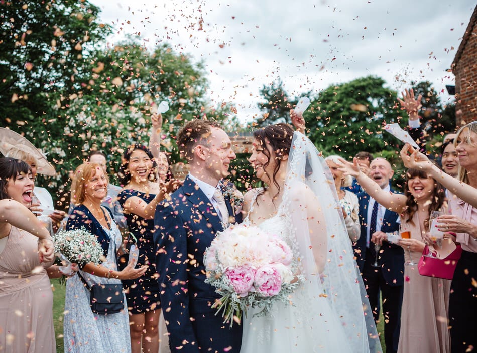 Bride and Groom face each other smiling as dried petal confetti is thrown over them by happy wedding guests