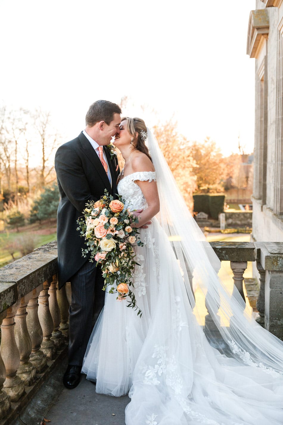 Bride and Groom share a kiss on the balcony at Bourton Hall. Bride wears floaty off the shoulder wedding dress, with full length veil