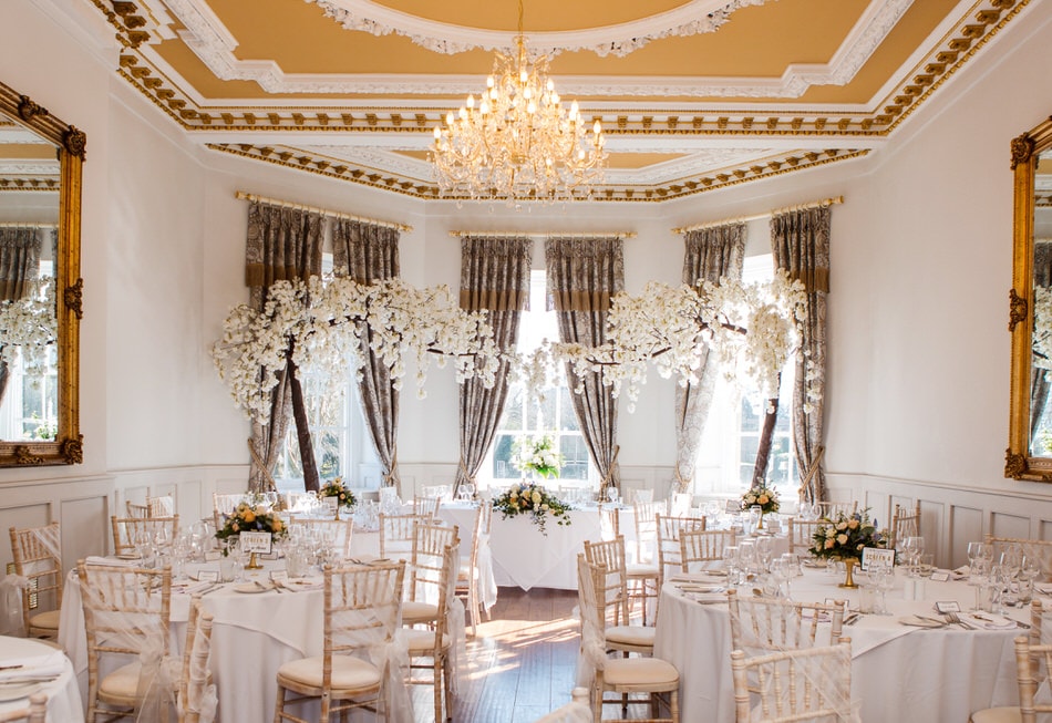 room with blossom trees set up for a wedding meal. White table linen and crystal chandelier