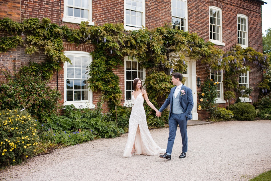 Groom wearing a mid blue suit with light blue waistcoat, walking hand in hand with his Bride, in front of a country house in the West Midlands, covered in foliage