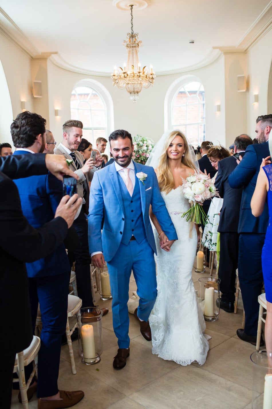 Smiling newly married Groom in bright blue suit holds hands with his Bride and walks down the aisle at Iscoyd Park