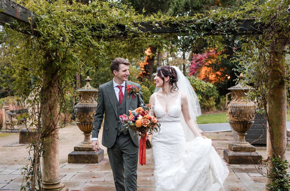 Bride holding a bouquet of Autumn coloured flowers with rust ribbons, holds her dress in one hand and walks under a foliage covered pergola, smiling at her Groom, after their Moxhull Hall wedding