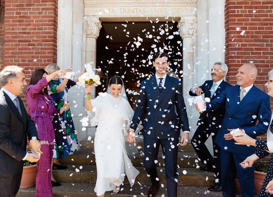 Bride wearing a mid length wedding dress holds her bouquet up high and walks out of Holy Trinity church, Sutton Coldfield with her Groom, through a blizzard of white paper confetti