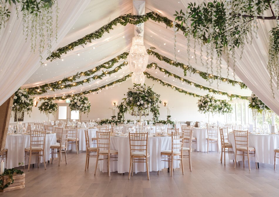 Interior of a wedding marquee styled with white ceiling drapes and hanging green foliage