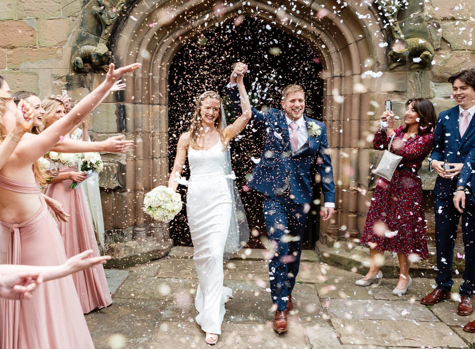 Just married Bride and Groom walk out of a Birmingham church with their hands raised above their heads. White and pink paper confetti is being thrown