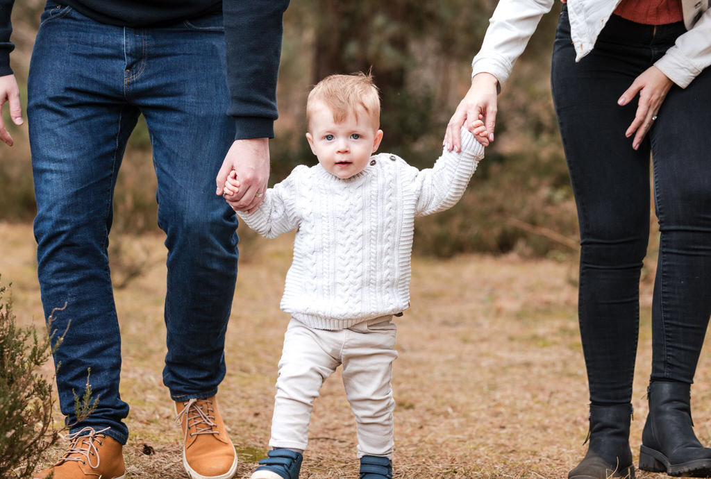 a young child holds his parents hands and looks at the camera in a natural setting of Sutton park