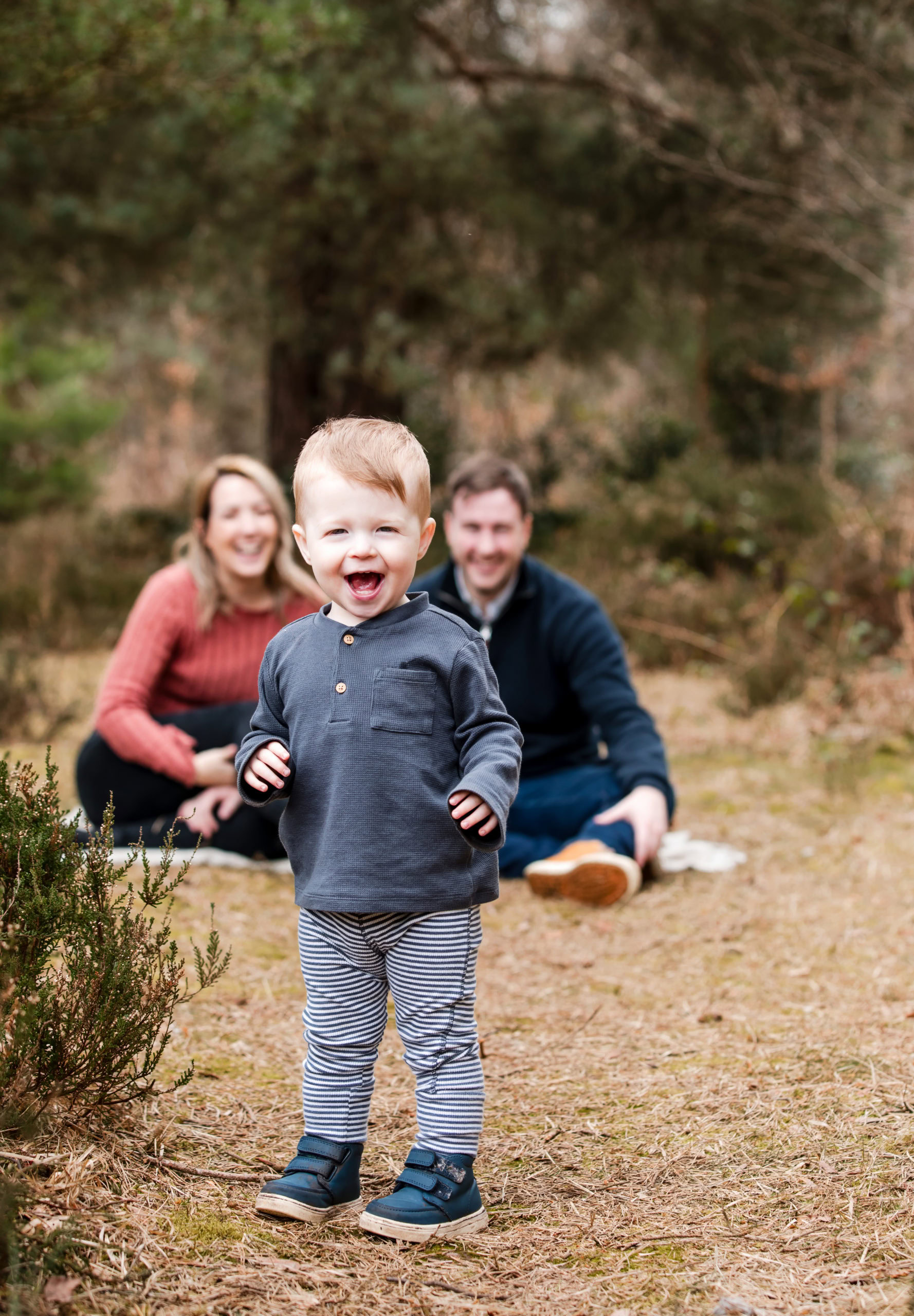 A toddler laughing and walking toward the camera during a relaxed family photoshoot in Sutton Park, with parents smiling in the background.