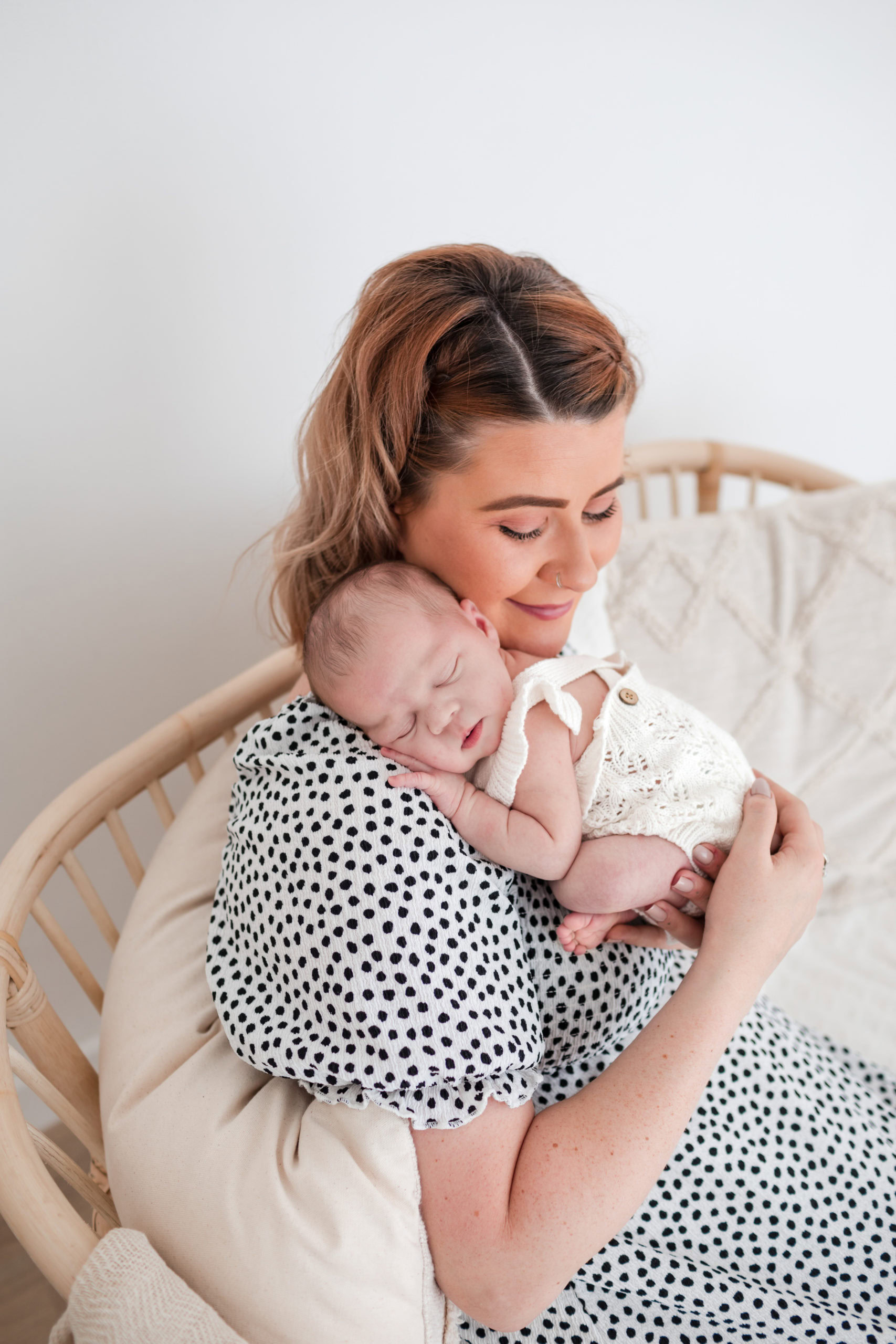 Woman holding a newborn baby asleep on her chest, sitting in a light, neutral room with a wicker chair and soft blankets