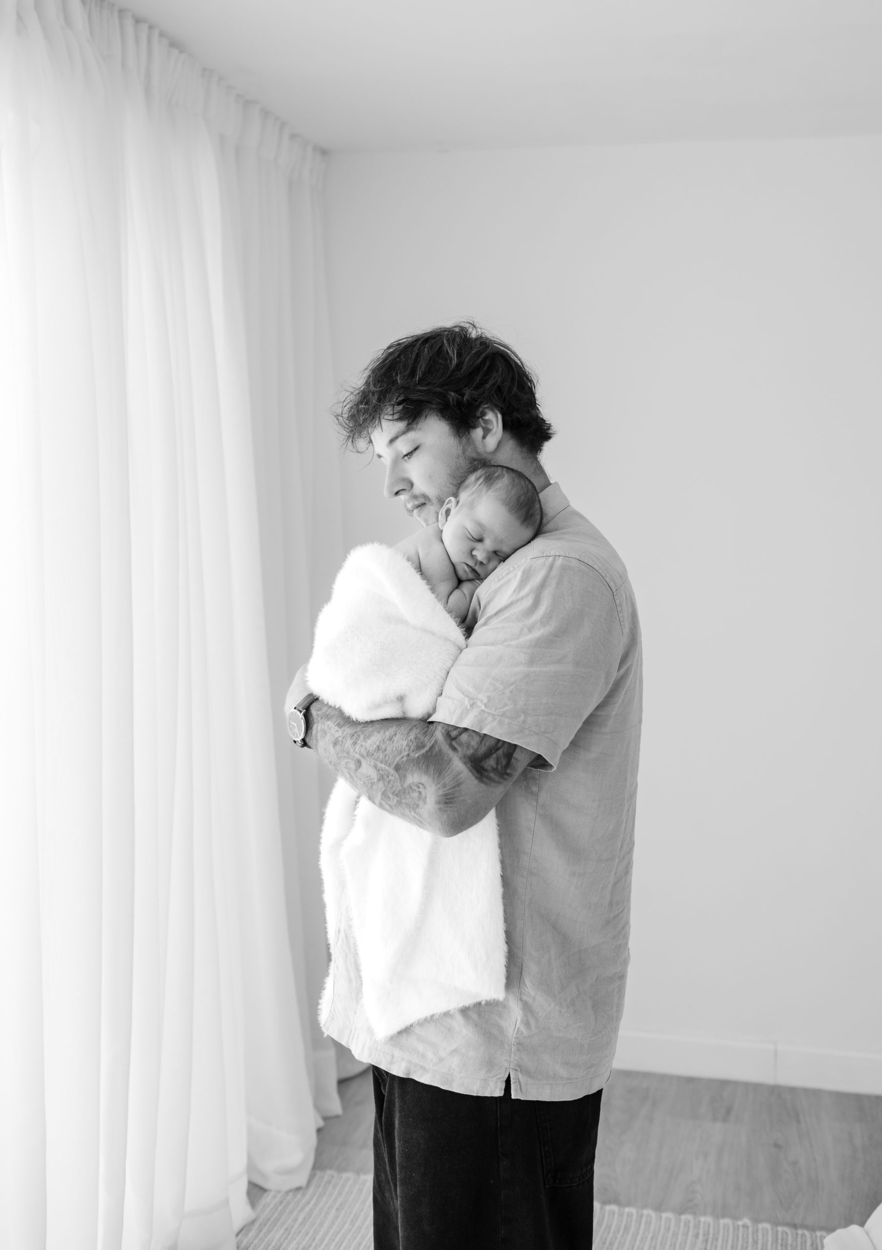 black and white image of Dad with Tattoo sleeve holding newborn baby wrapped in a fluffy blanket on his chest