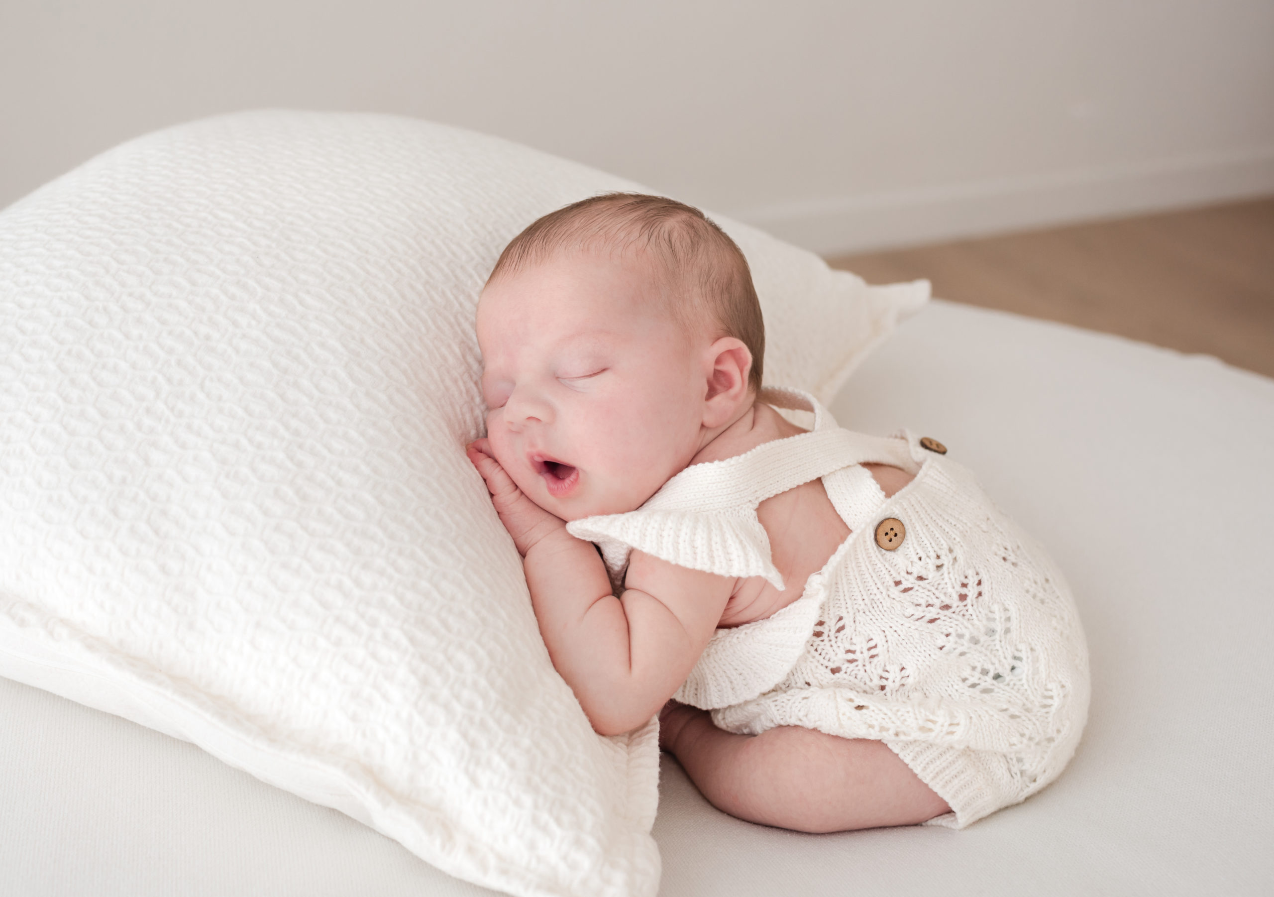 newborn baby girl asleep on a cream cushion wearing a strappy cream knitted romper