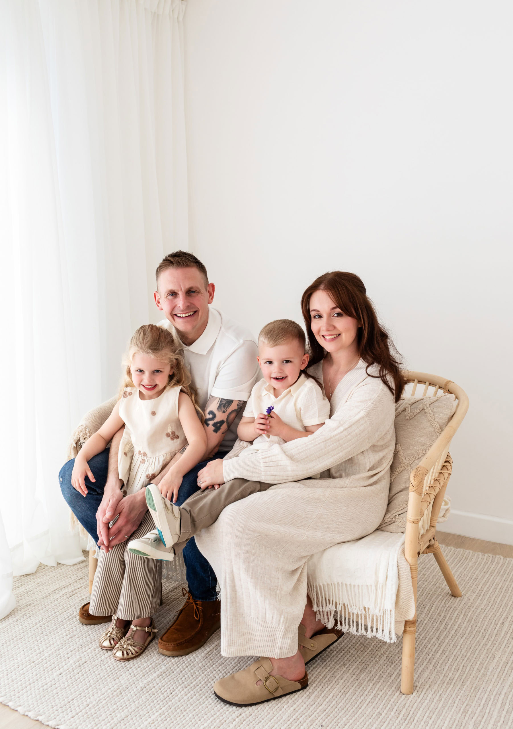 Mum, Dad and two young children, wearing light coloured clothes sit on a wicker sofa and cuddle for a photo