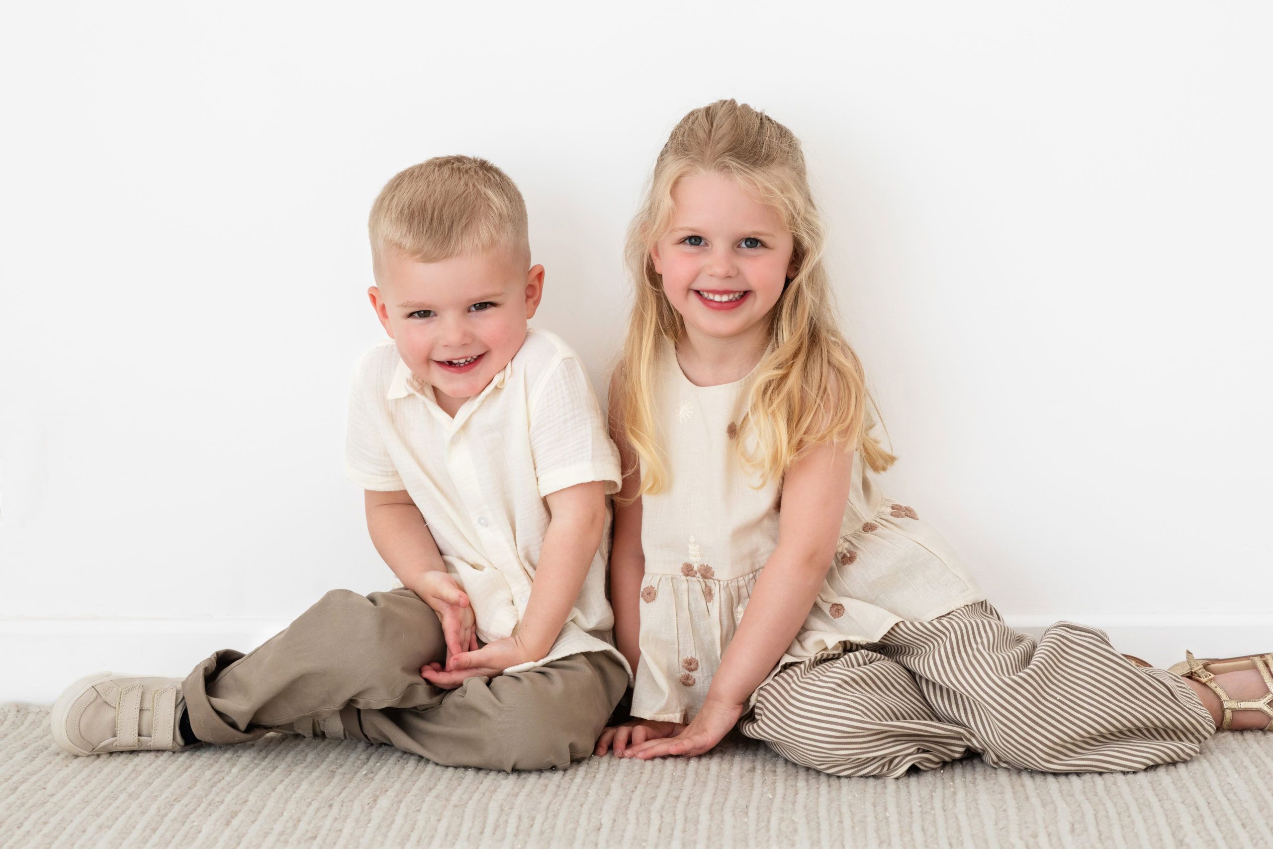 A young girl and boy wearing cream and beige coloured clothes sitting on a rug in a light photography studio in Sutton Coldfield