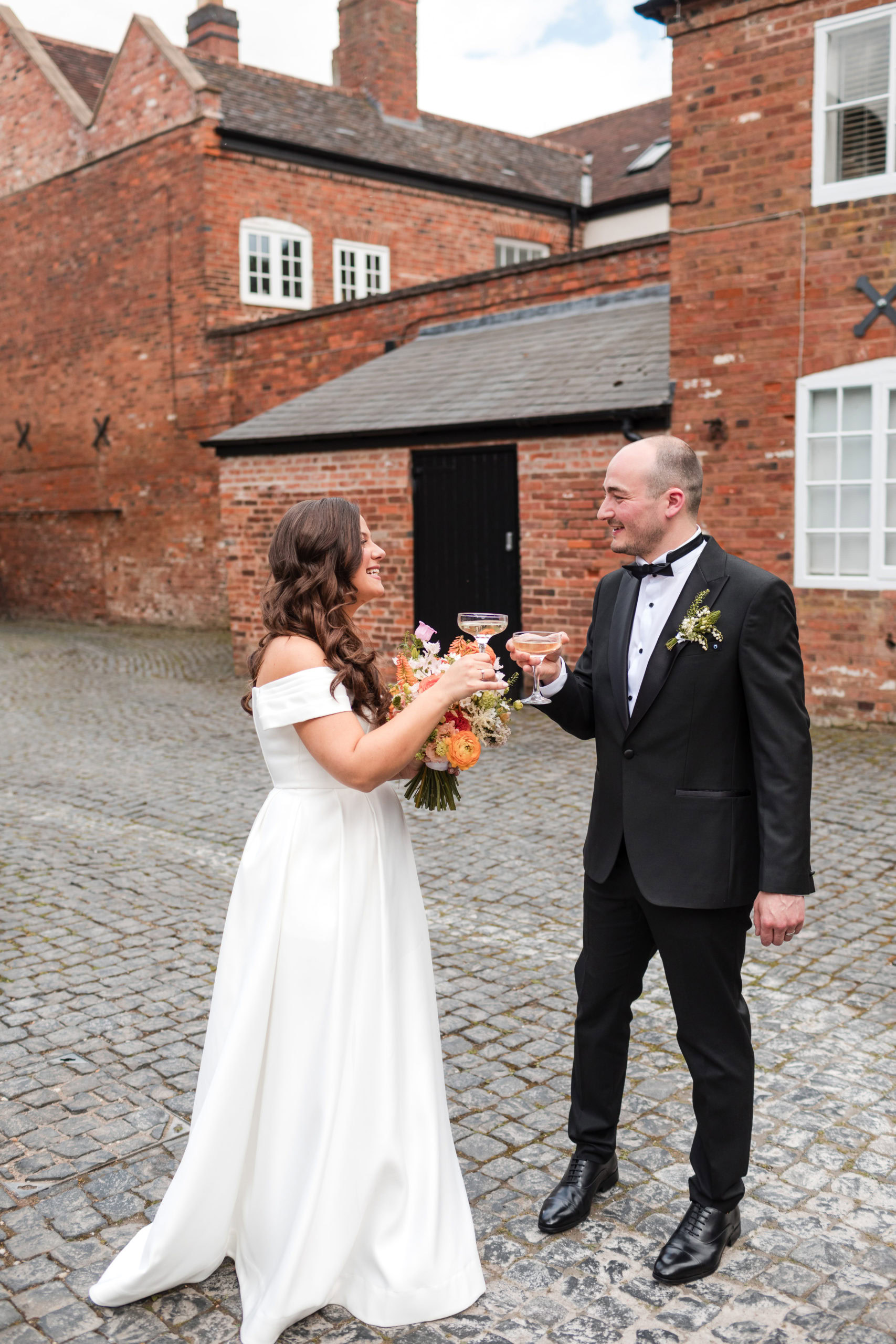 Candid moment of a Bride and Groom wearing a tuxedo holding champagne glasses and a colourful bouquet